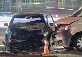 Damaged in heavy car accident vehicles after collision on city street crash site at night.